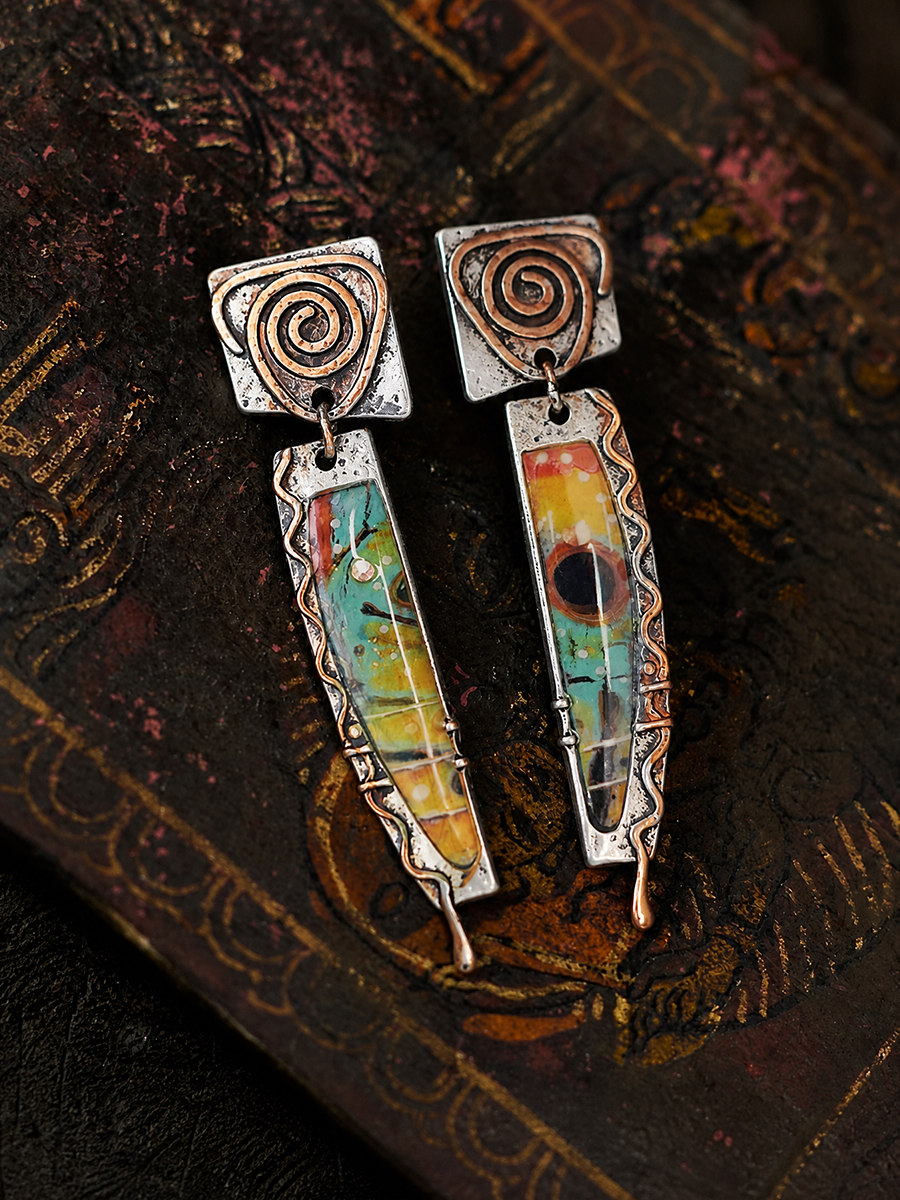 A pair of handmade dangle earrings with oxidized silver spiral tops and colorful inlay panels featuring green, yellow, orange, and red, displayed on a dark textured book.