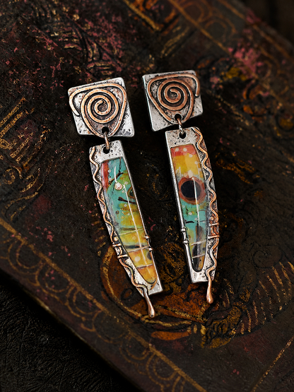 A pair of handmade dangle earrings with oxidized silver spiral tops and colorful inlay panels featuring green, yellow, orange, and red, displayed on a dark textured book.