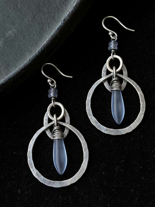A pair of silver dangle earrings with hammered circular wire frames and teardrop-shaped blue-gray beads, displayed on a black background.