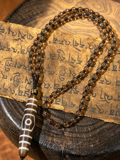A beaded necklace with dark brown translucent beads and a large white-and-brown striped cylindrical pendant, displayed on a rustic wooden surface with an aged paper featuring script.