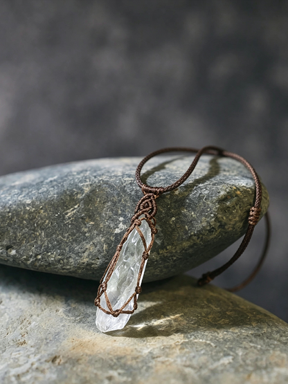 A clear quartz crystal pendant necklace wrapped in a rustic brown cord, resting on a smooth gray stone against a dark, textured gray background.