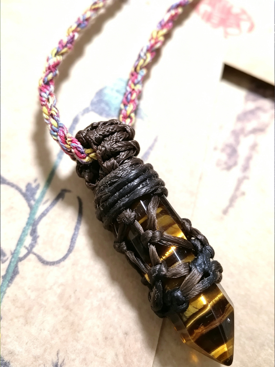 A handmade pendant necklace featuring a pointed tiger's eye crystal wrapped in dark brown cord, suspended from a braided multicolor cord with pink, yellow, and blue threads, resting on a vintage-style paper background.