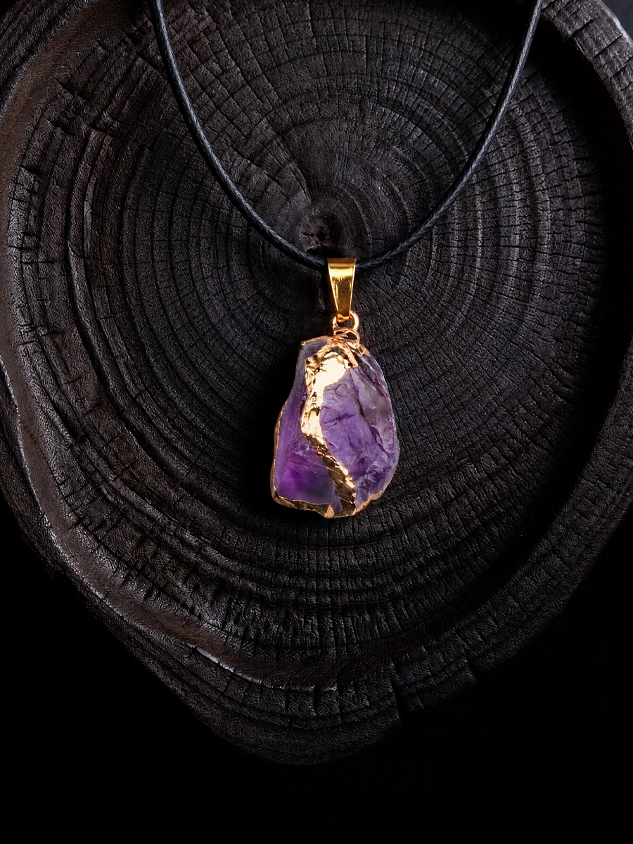 A raw amethyst crystal pendant necklace with a gold bail and black cord, displayed on a dark wood slice background.