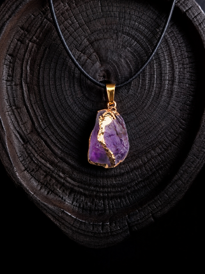 A raw amethyst crystal pendant necklace with a gold bail and black cord, displayed on a dark wood slice background.