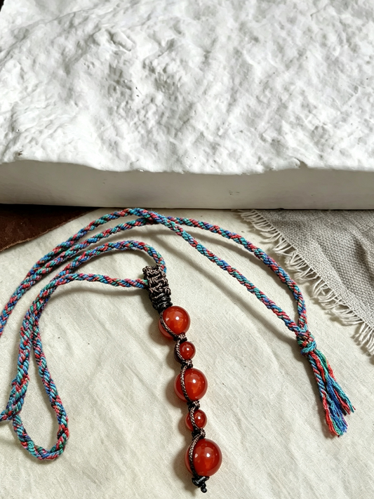 A handmade beaded necklace with four red-orange carnelian beads threaded on a blue, red, and white braided cord, displayed on a textured white fabric surface.