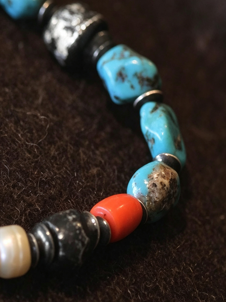A close-up of a beaded necklace featuring turquoise-blue marbled beads, a bright coral-red bead, a white pearl-like bead, and dark metallic spacer beads on a dark brown textured background.