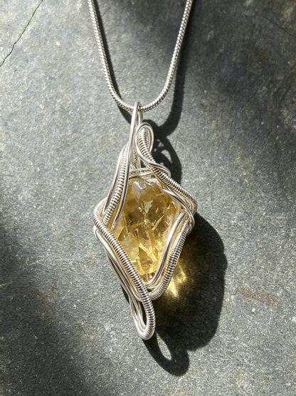 A handmade silver wire-wrapped pendant necklace featuring a rough, faceted yellow gemstone, displayed on a dark textured stone surface with dramatic natural sunlight and shadows.
