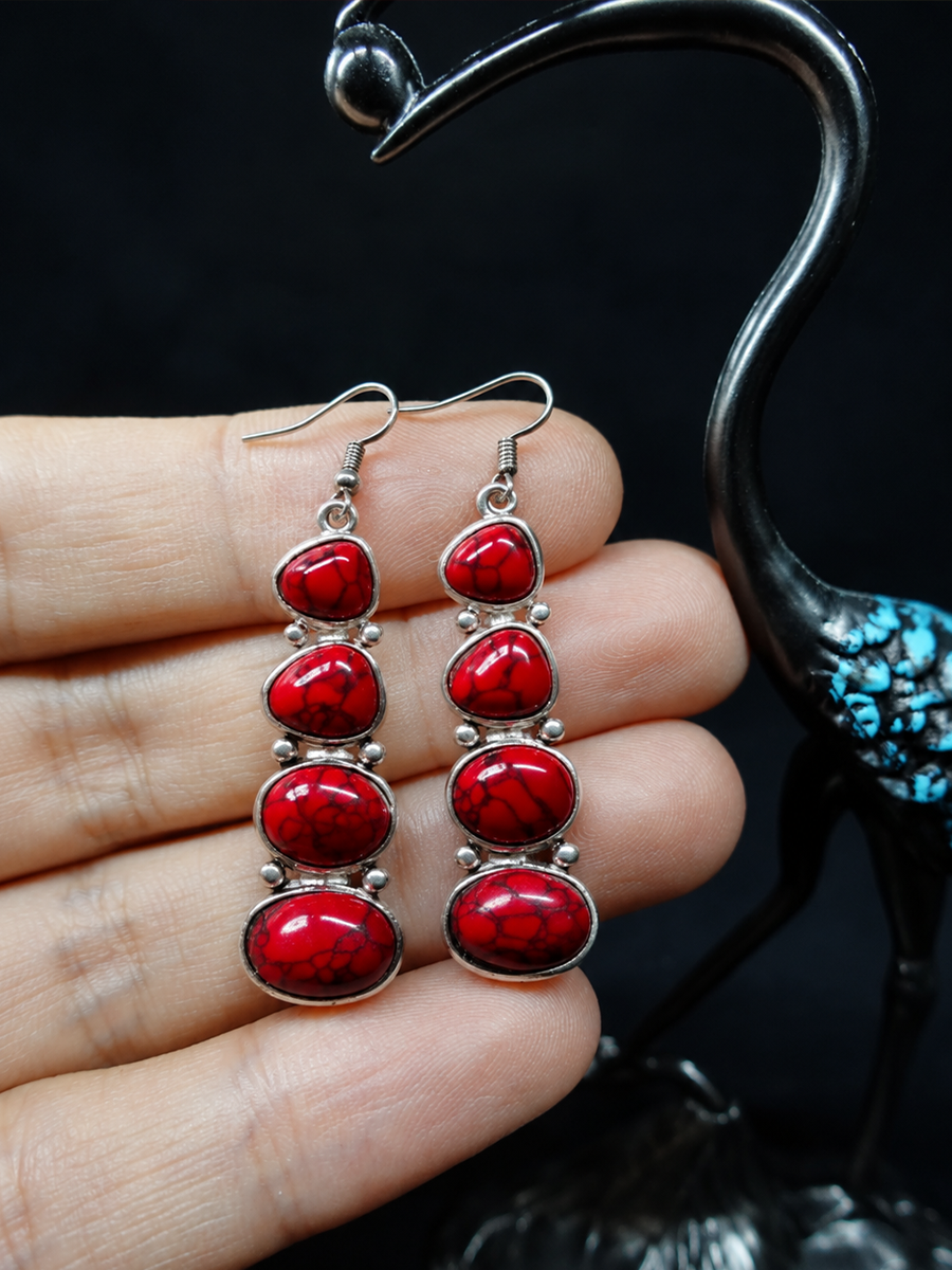 A pair of dangling earrings with oval red stones set in silver-tone metal, held between fingers against a dark background with a decorative black bird sculpture.