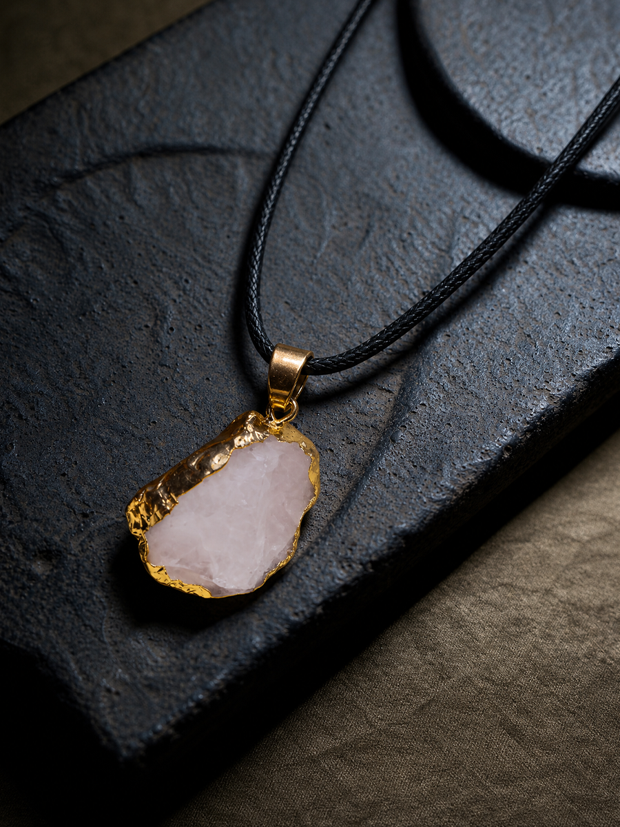 A raw, irregularly shaped pale pink stone pendant with a hammered gold-tone rim, suspended on a black cord necklace, displayed on a dark textured slate surface.