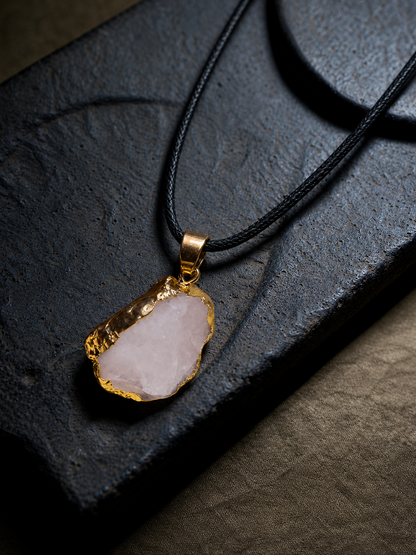 A raw, irregularly shaped pale pink stone pendant with a hammered gold-tone rim, suspended on a black cord necklace, displayed on a dark textured slate surface.