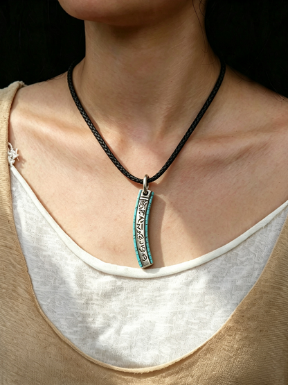 A close-up of a person wearing a black braided cord necklace with a vertical silver-toned pendant featuring turquoise blue inlay and engraved details.