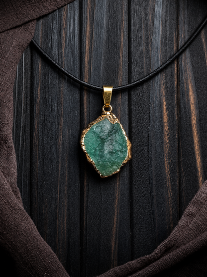 A raw green stone pendant with a gold-toned bezel setting, hanging from a black cord necklace against a dark wood background with draped brown fabric.