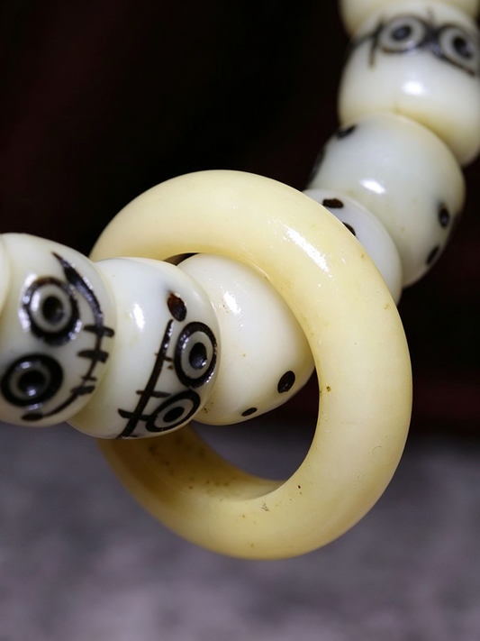 A close-up of a cream-colored beaded necklace or bracelet featuring a large smooth ring bead intertwined with round beads decorated with black hand-drawn face designs.