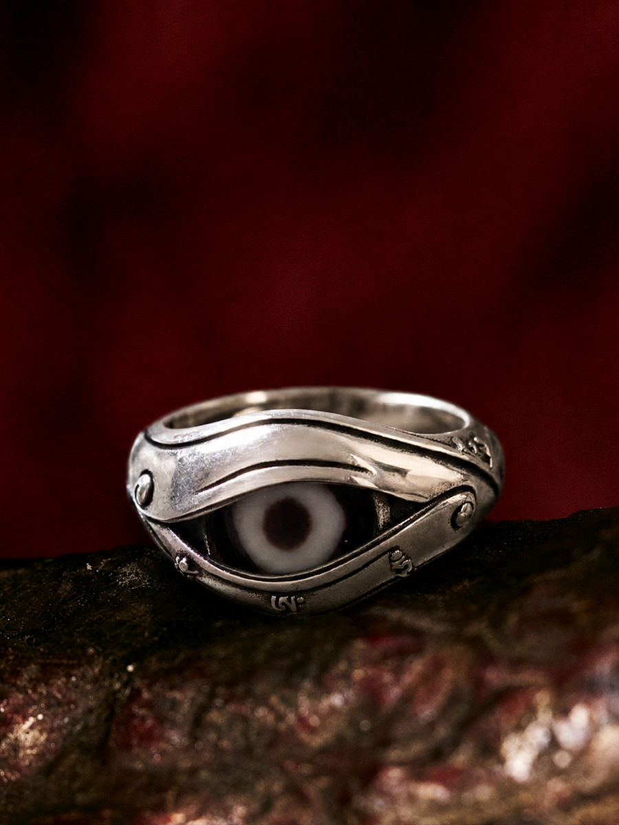 A silver-toned evil eye ring with a white and black glass eye inlay, resting on a dark textured surface against a dark red background.