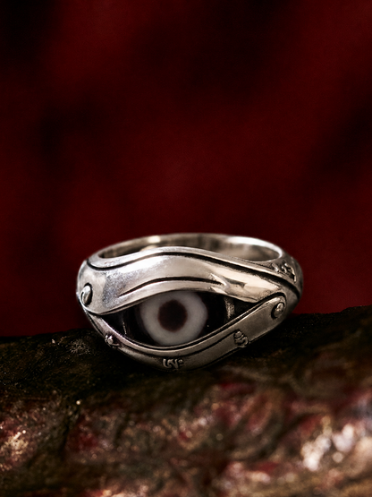 A silver-toned evil eye ring with a white and black glass eye inlay, resting on a dark textured surface against a dark red background.