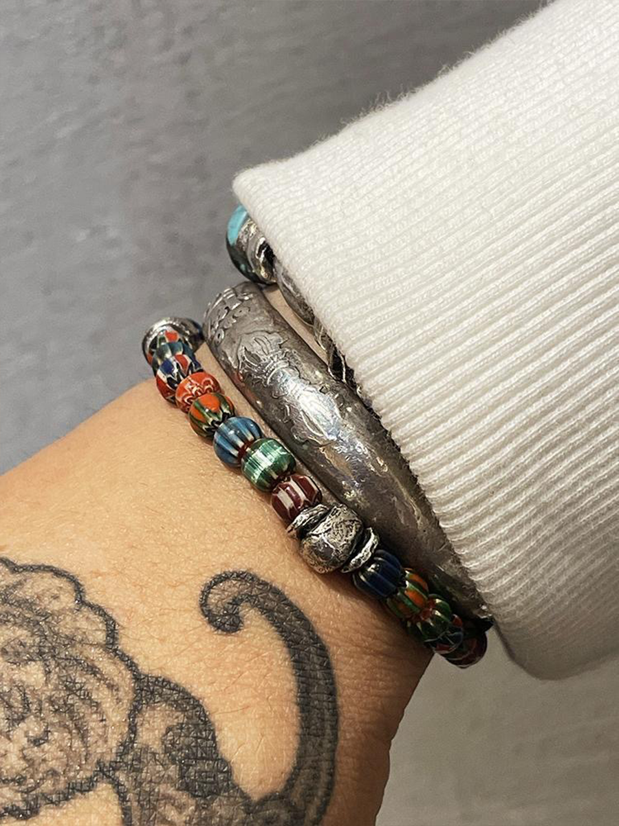 A close-up of a person's wrist wearing multiple bracelets, including a silver-toned engraved cuff, a beaded bracelet with red, blue, green, and brown beads, and a thin silver chain bracelet, next to a white ribbed sleeve.