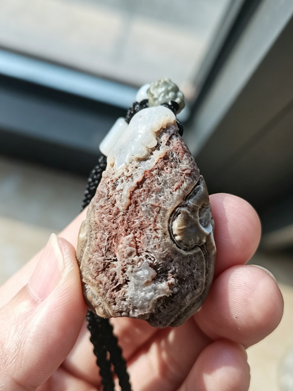 A person's hand holding a rough, textured teardrop-shaped stone pendant with a carved eye detail and a black cord, photographed in natural light near a window.