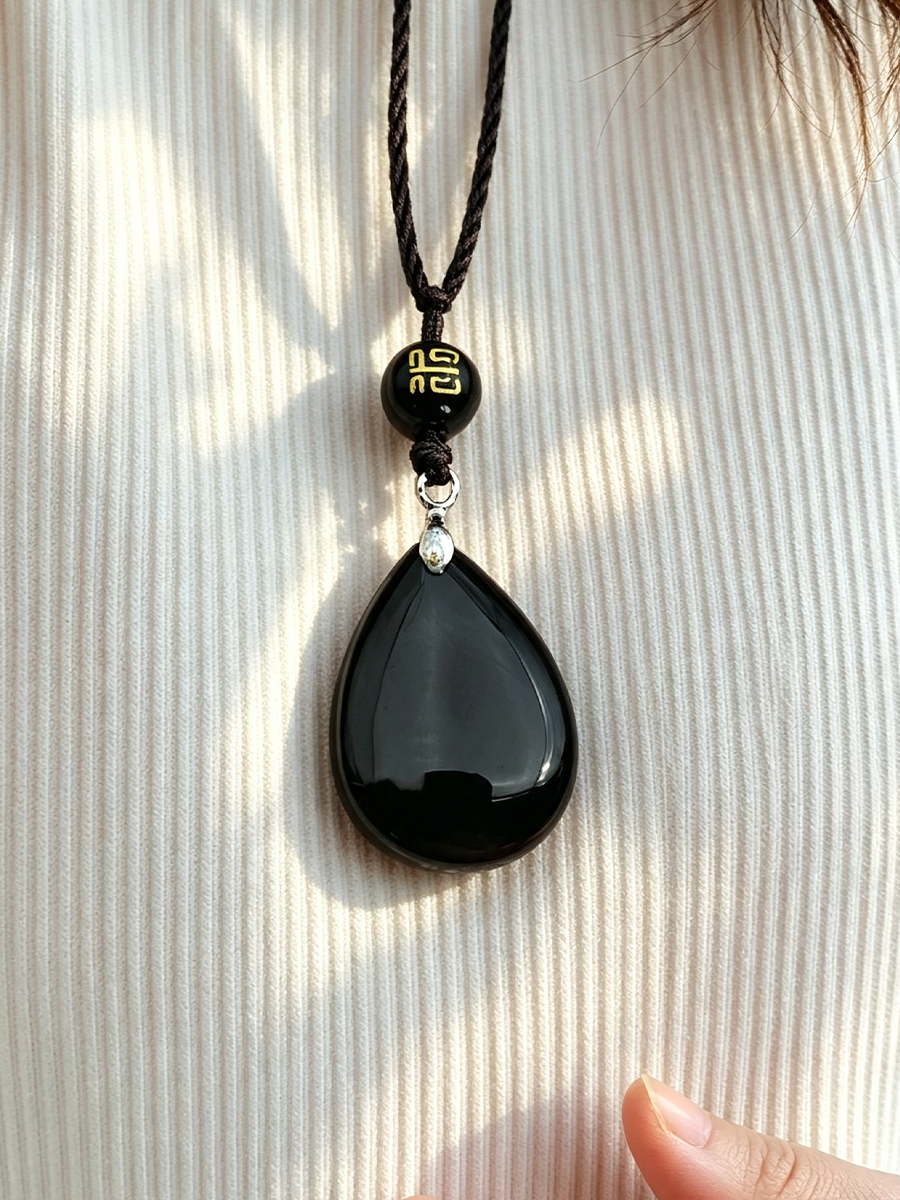 A close-up of a black teardrop pendant necklace with a black cord, featuring a small black bead with a gold Chinese character and a tiny silver spacer, worn against a ribbed cream shirt.