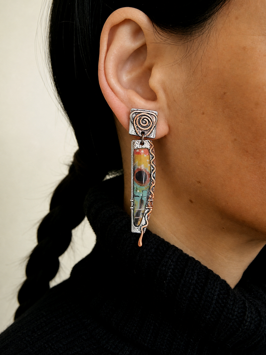 A close-up of a person's ear wearing a long, multi-layered dangle earring. The earring features a silver-toned spiral stud, a rectangular section with a colorful marbled pattern in yellow, orange, green, and blue, and a dangling textured silver bar ending in a small copper-colored bead.