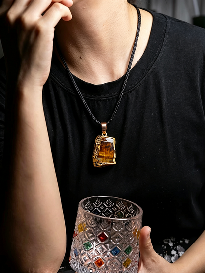 A person wearing a black t-shirt and a gold pendant necklace with a rough-cut amber stone, holding a clear patterned glass tumbler with colorful gem accents.