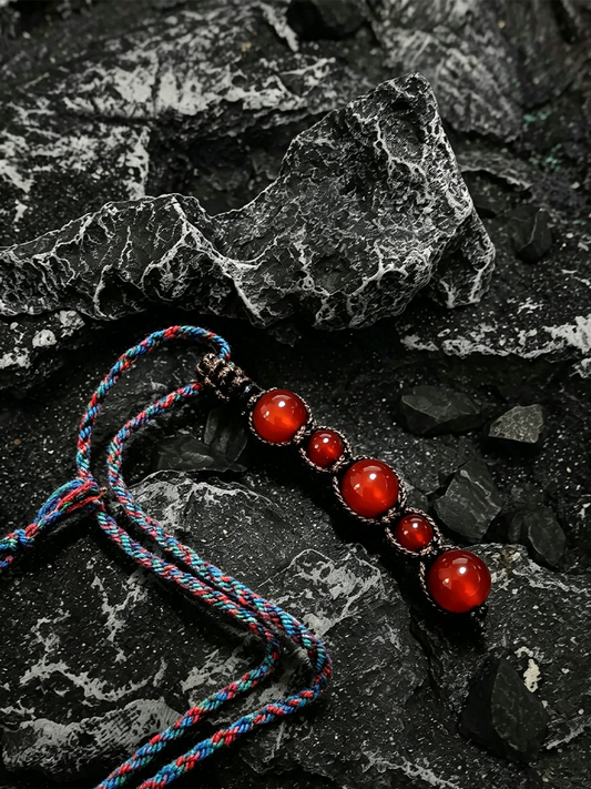 A beaded necklace with five glossy red cabochon beads on a braided blue, red, and black cord, displayed on a dark textured rock surface.