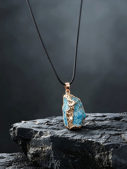 A raw turquoise-blue stone pendant necklace with gold-toned metal edging, hanging from a black cord on a dark textured rock against a dark gray background.