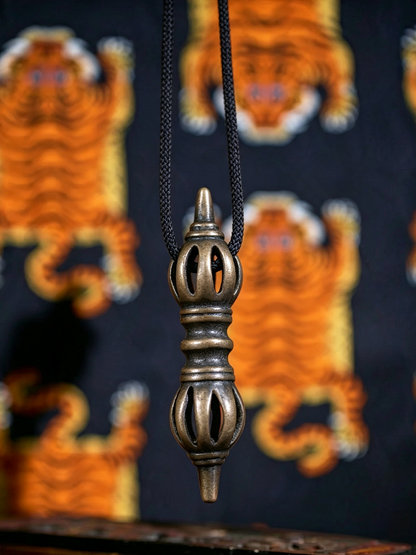 A bronze-toned metal pendant with a pointed top and decorative cutouts, hanging from a black cord against a dark fabric background with orange tiger motifs.