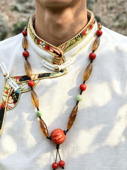 A close-up of a person wearing a traditional white embroidered Tibetan-style collar with a long beaded necklace featuring amber-brown elongated beads, red round beads, and small light green beads.