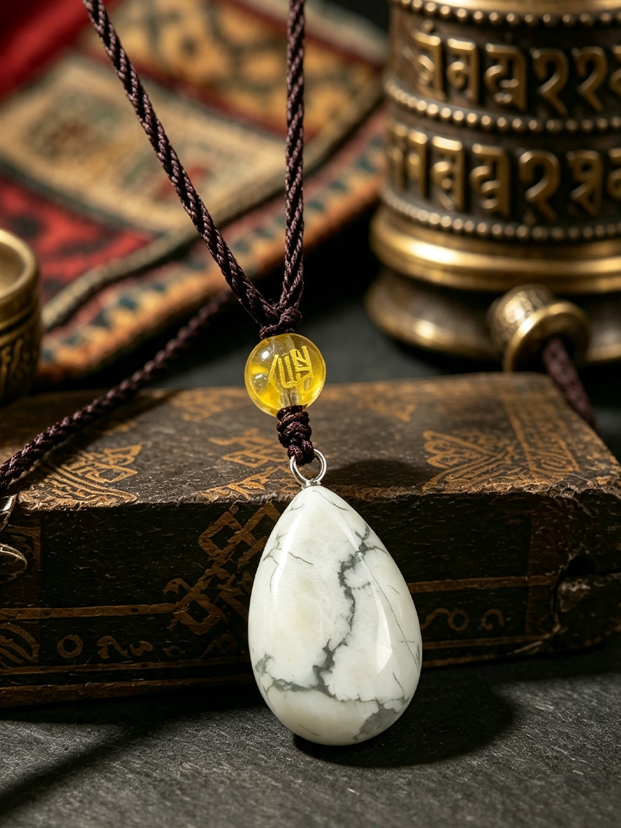A teardrop-shaped white marble pendant necklace with dark brown braided cord and a small yellow translucent bead, displayed on a dark surface with vintage books and a brass prayer wheel in the background.