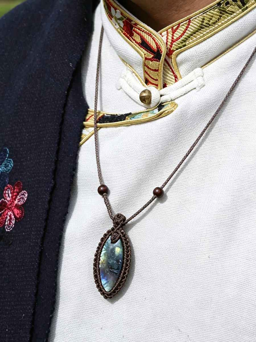 A close-up of a person wearing a dark brown cord necklace with a marquis-shaped iridescent abalone shell pendant, styled over a white embroidered traditional shirt and a navy cardigan.