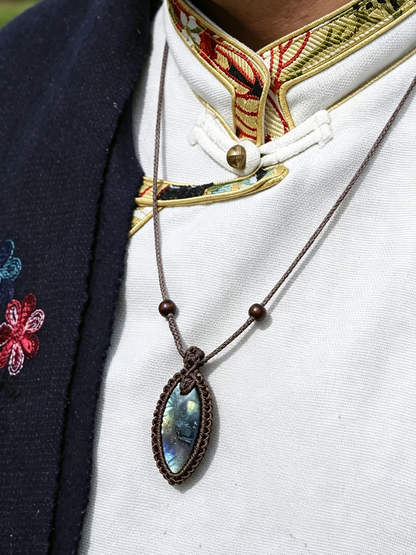 A close-up of a person wearing a dark brown cord necklace with a marquis-shaped iridescent abalone shell pendant, styled over a white embroidered traditional shirt and a navy cardigan.