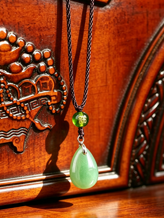 A green jadeite teardrop pendant necklace with a black braided cord and a small green bead featuring Chinese characters, displayed against a carved wooden background.