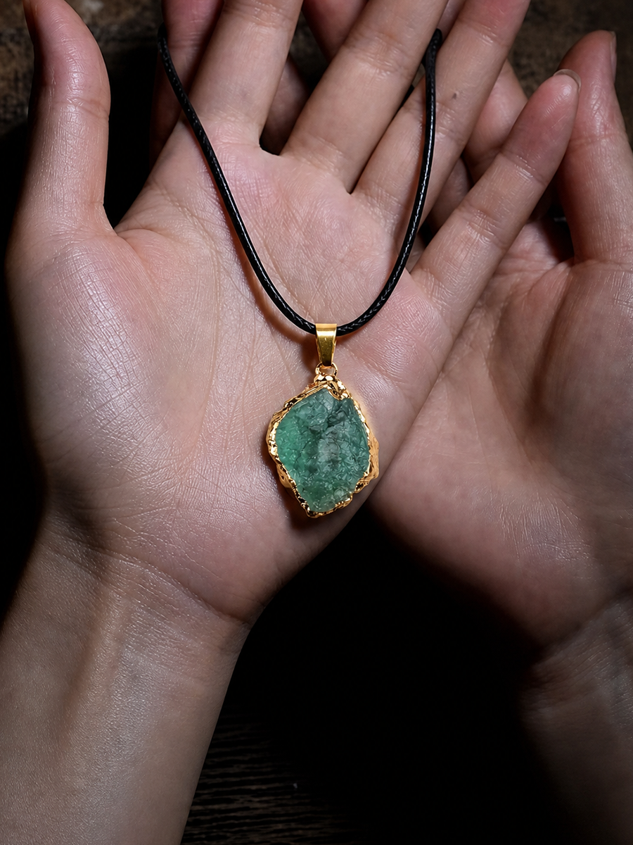 A handmade pendant necklace featuring a raw green stone with a gold bezel setting, held in two hands against a dark background.