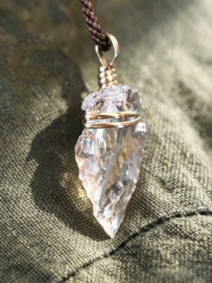 A natural clear quartz crystal point pendant wrapped in silver-toned wire, hanging from a dark brown cord, set against a textured olive green fabric background.