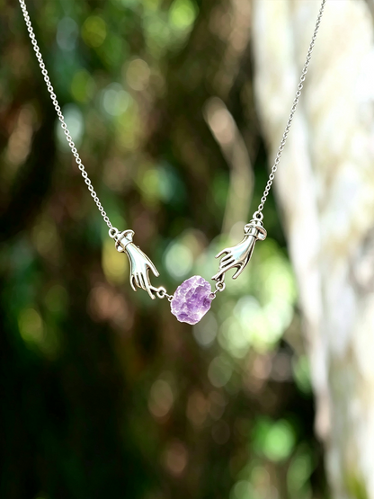 A silver necklace featuring two hands reaching toward a small, faceted purple amethyst gemstone, photographed against a blurred natural outdoor background of green foliage and light wood.