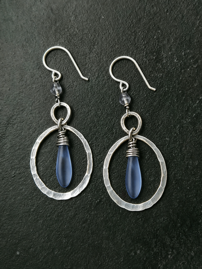 A pair of handmade dangle earrings featuring hammered silver-tone circular frames, small clear faceted beads, and teardrop-shaped blue glass drops, displayed on a dark textured slate background.