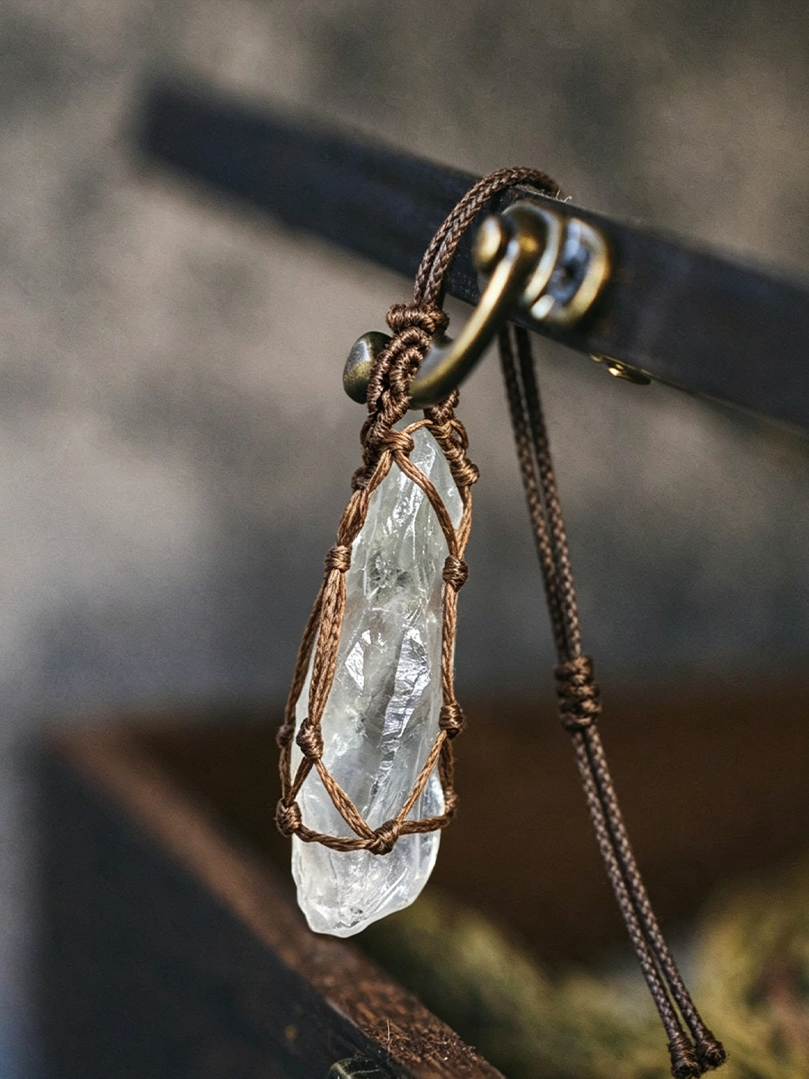 A clear quartz crystal pendant wrapped in a brown macrame cord and hanging from a dark brown braided necklace cord with a brass-tone ring clasp.