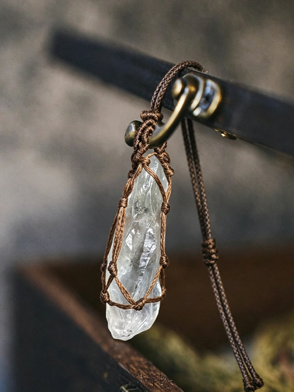 A clear quartz crystal pendant wrapped in a brown macrame cord and hanging from a dark brown braided necklace cord with a brass-tone ring clasp.