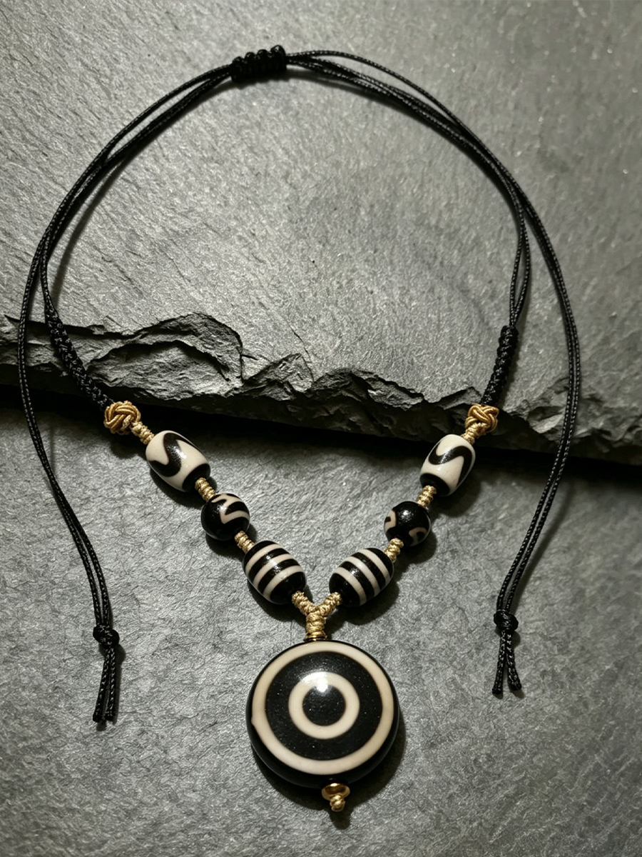 A black cord necklace with a circular evil eye pendant and striped beads, displayed on a textured gray slate surface.
