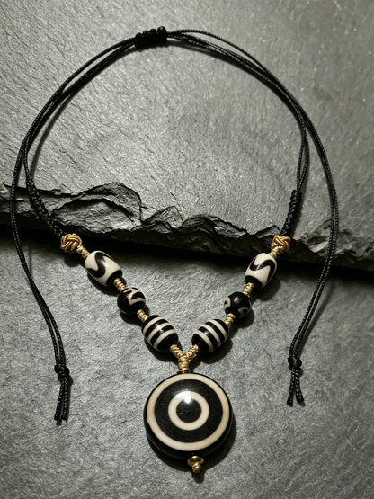 A black cord necklace with a circular evil eye pendant and striped beads, displayed on a textured gray slate surface.