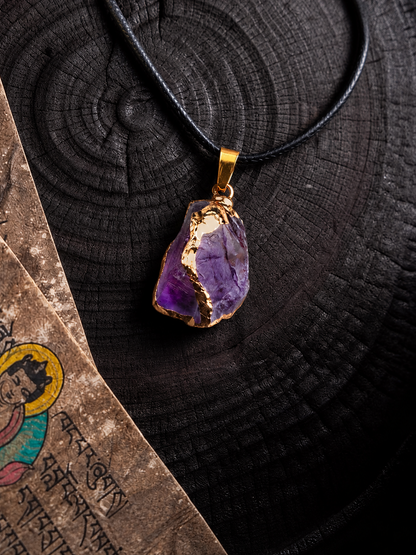A raw amethyst pendant necklace with a gold-toned bezel setting and black cord, displayed on a dark wood slice background next to an old parchment scroll.