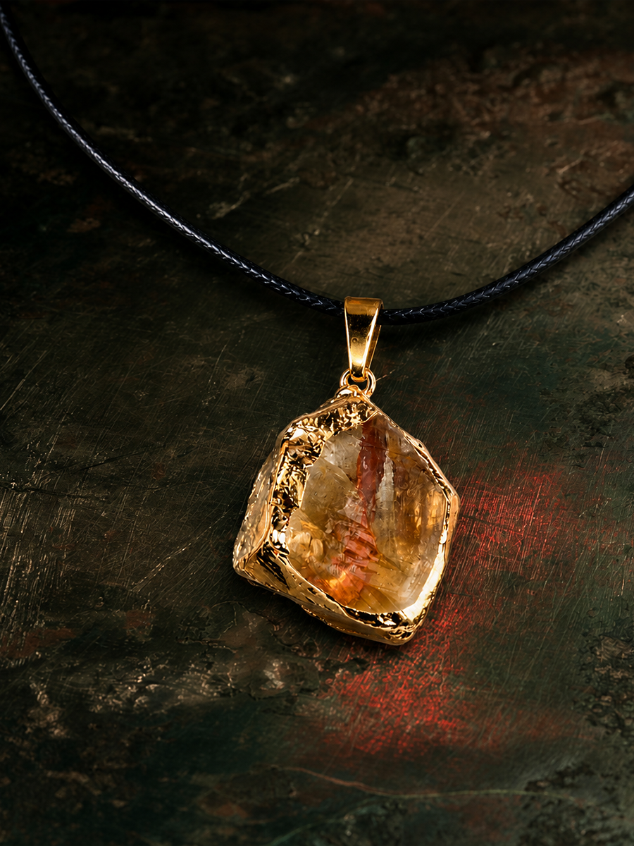 A gold pendant necklace featuring a raw, irregular faceted amber gemstone, suspended from a black cord against a dark textured background.