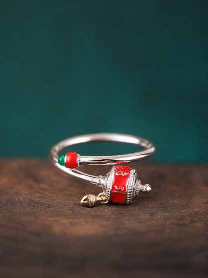 A silver-toned bangle bracelet with a red and white patterned cylindrical charm, a small red bead, and a tiny gold bell, displayed on a dark wooden surface against a dark green background.