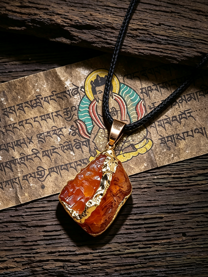 A raw amber stone pendant necklace with gold trim and a black cord, displayed on a textured wooden surface next to a printed paper with Tibetan script and a deity illustration.