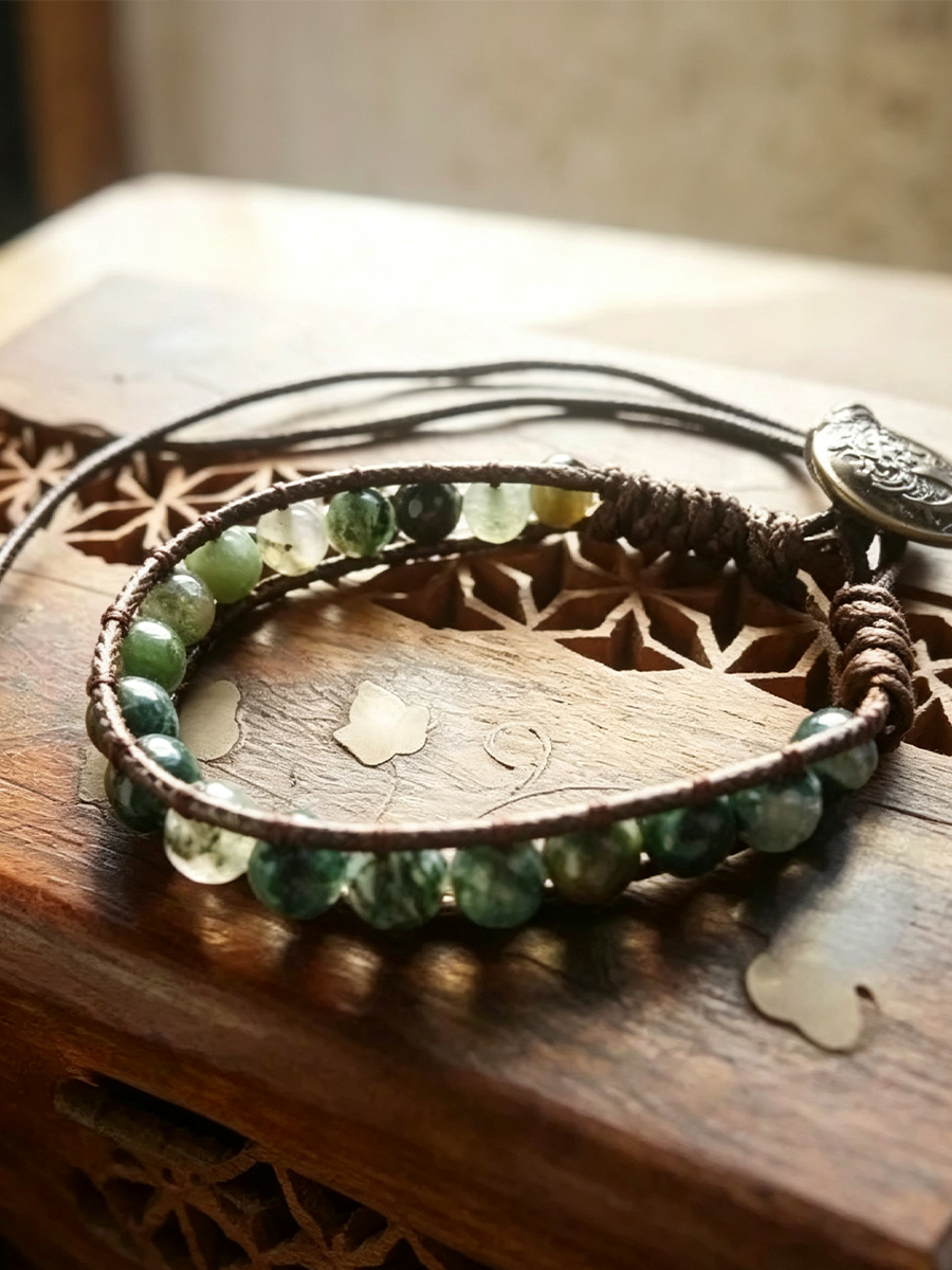 A handmade multi-strand bracelet featuring dark brown cords, polished green marbled beads, and a silver-toned engraved charm, displayed on a rustic wooden surface.