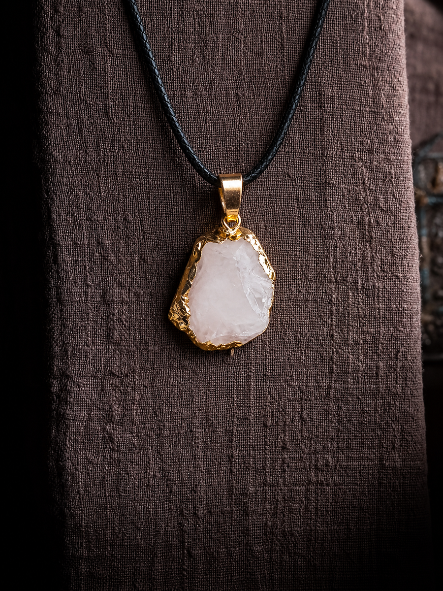 A raw, irregular-shaped white stone pendant with a gold bezel setting, hanging from a black cord necklace against a dark brown textured fabric background.