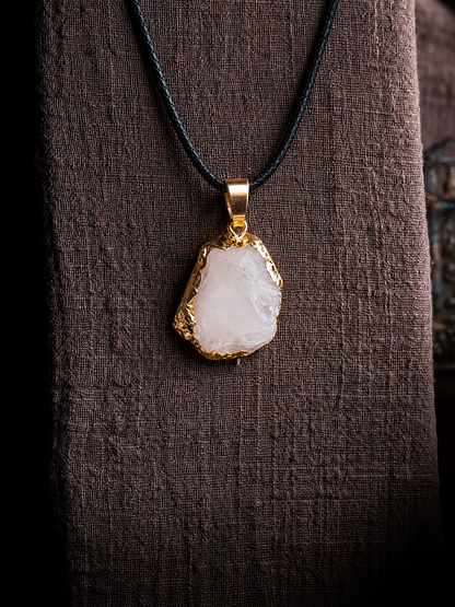 A raw, irregular-shaped white stone pendant with a gold bezel setting, hanging from a black cord necklace against a dark brown textured fabric background.