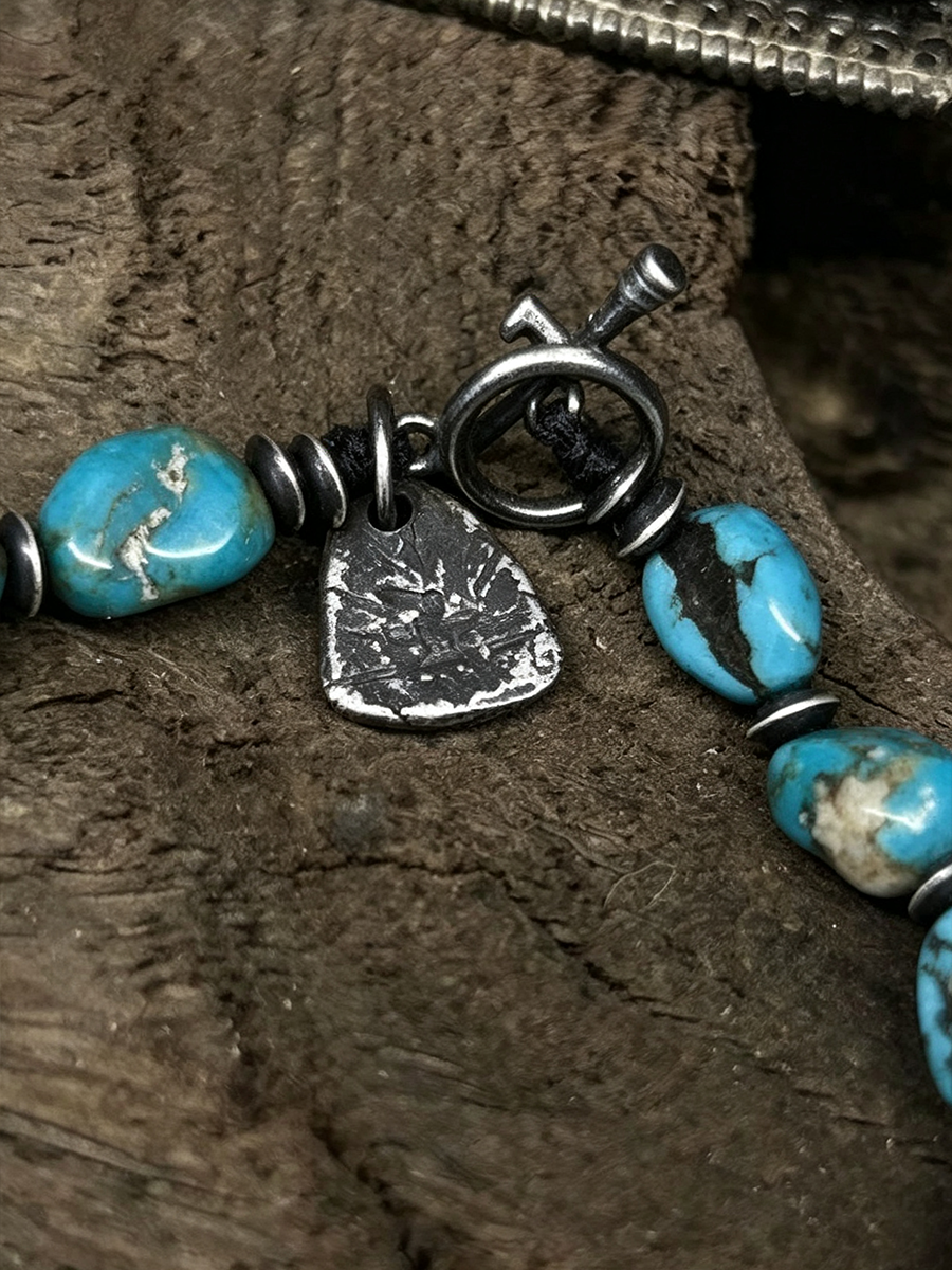 Close-up of a beaded jewelry strand with turquoise-blue marbled beads, dark metal spacers, a silver-toned engraved charm, and a toggle clasp resting on a textured brown wood surface.