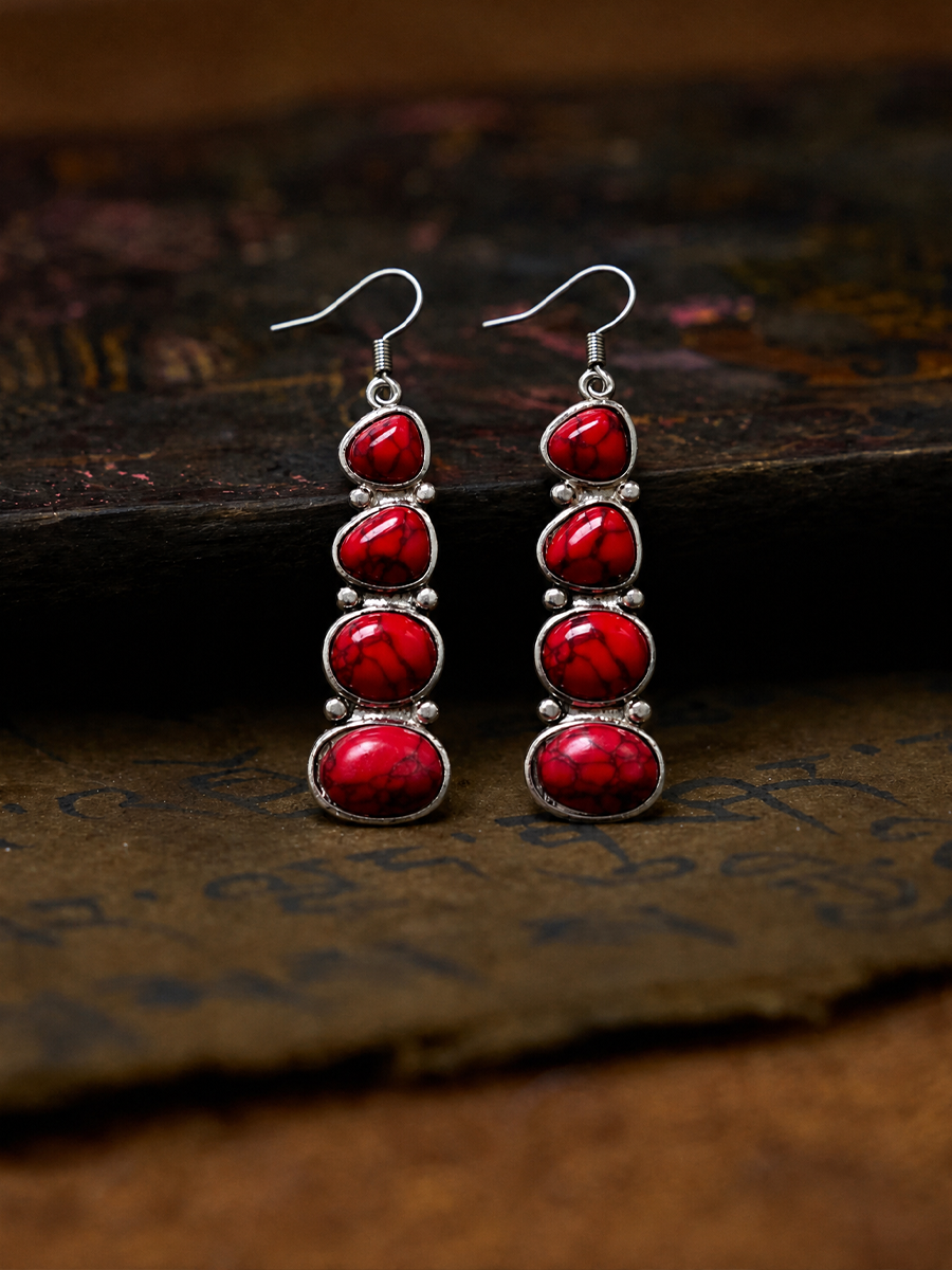 A pair of dangle earrings with four stacked red cabochon stones in silver bezel settings, hanging from silver fishhook wires. The earrings are displayed on a textured, dark brown surface.