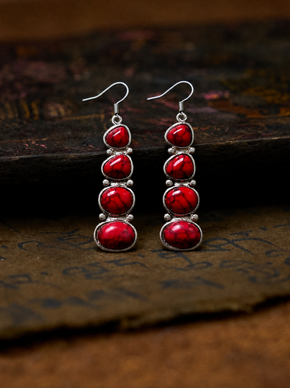 A pair of dangle earrings with four stacked red cabochon stones in silver bezel settings, hanging from silver fishhook wires. The earrings are displayed on a textured, dark brown surface.